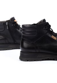 Des bottines en cuir noir présentent une surface texturée, un laçage à l'avant et une fermeture éclair sur le côté. La semelle est robuste avec un léger talon pour un meilleur soutien.