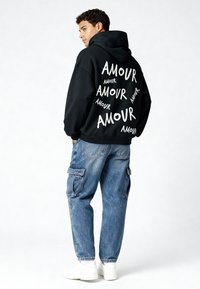 Felpa nera con cappuccio caratterizzata da testo bianco ripetuto "AMOUR," abbinata a jeans cargo blu chiaro e sneakers bianche. Modello mostrato di spalle.