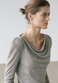 Jeune femme aux cheveux attachés, portant un haut gris à manches longues avec un col drapé, tournée vers la droite devant un fond uni.