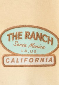 Écusson brodé avec le texte : « THE RANCH Santa Monica LA, US » dans un ovale bleu et « CALIFORNIA » dans un rectangle sur tissu beige.