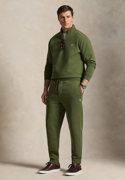 Férfi, olíva zöld pulóvert és hozzáillő melegítőnadrágot visel, barna sportcipő, napszemüveg lóg a nyakkivágásról, és karóra, semleges fal előtt áll.