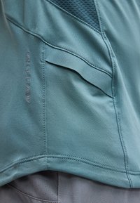 Κοντινή λήψη τιρκουάζ αθλητικού υφάσματος με τσέπη, ορατές ραφές και κείμενο «DRI-FIT ADV», φορεμένο πάνω από διάτρητο γκρι υλικό.