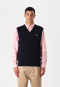 Ναυτικό πλεκτό γιλέκο με σχέδιο καλωδίου, σχεδίαση v-neck, φορεμένο πάνω από ένα ανοιχτό ροζ πουκάμισο με γιακά, συνδυασμένο με καφέ παντελόνι. Παρών το λογότυπο Lacoste.