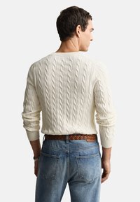 Roomkleurige kabelgebreide trui met lange mouwen, geribbelde manchetten en een ronde halslijn, gecombineerd met lichtblauwe spijkerbroek en een gevlochten bruine riem.