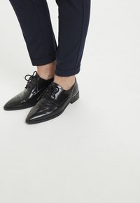 Chaussures de ville en cuir noir à bout pointu et à lacets, associées à un pantalon bleu marine fuselé avec un ourlet à la cheville.