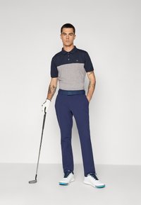Marineblå og grå kortermet poloskjorte med krage, sammen med marineblå bukser. Mannen holder en golfkølle, iført hvite og blå golf sko.