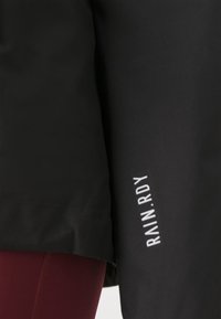 Black rain jacket with a smooth texture, featuring a white "RAIN.RDY" logo on the side, paired with burgundy leggings underneath.