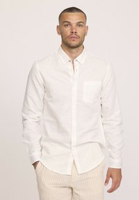 Chemise boutonnée en lin blanc avec une poche poitrine, manches longues et un ourlet légèrement arrondi, accompagnée de pantalons à rayures claires.