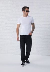 Weißes, kurzärmliges, eng anliegendes T-Shirt kombiniert mit dunklen Jeans und schwarzen Sneakers. Das Model trägt schwarze Sonnenbrillen und zeigt ein Tattoo auf seinem Arm.