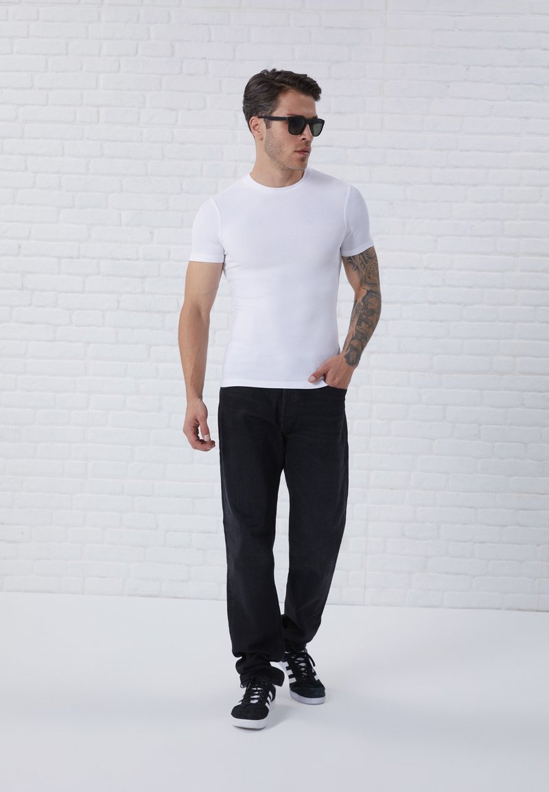Weißes, kurzärmliges, eng anliegendes T-Shirt kombiniert mit dunklen Jeans und schwarzen Sneakers. Das Model trägt schwarze Sonnenbrillen und zeigt ein Tattoo auf seinem Arm.