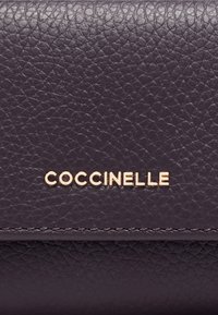Superficie in pelle trama scura con il nome del marchio "COCCINELLE" in metallo dorato centrato sopra la cucitura.