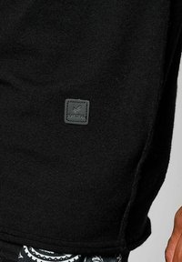 Tessuto a maglia nero con una superficie testurizzata, caratterizzato da un'etichetta quadrata in pelle nera con logo "Kaporal" in rilievo e accenti diagonali.