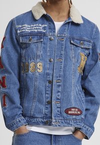 Denim jacket with shearling collar, blue color, embroidered text, golden and red patches, button-up front, and two chest pockets.