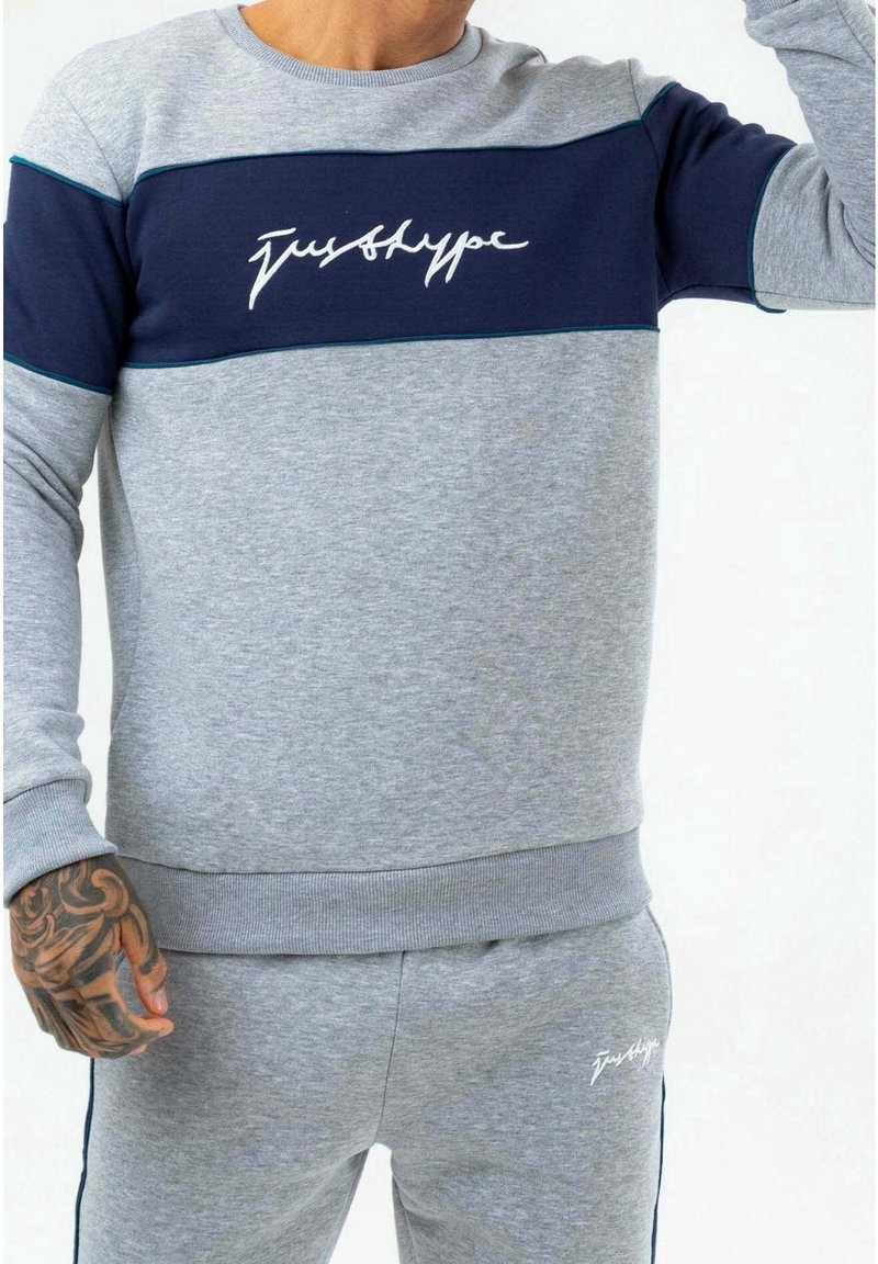 Szary sweter z granatowym akcentem, z białym logo. Żebrowane mankiety i dół, gładka tekstura oraz luźny krój, prezentowany z pasującymi spodniami.