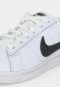 Λευκό αθλητικό παπούτσι Nike με μαύρο λογότυπο swoosh, λευκά κορδόνια και υφή στη σόλα σε ανοιχτό γκρι φόντο.