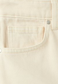 Tissu en denim de couleur crème montrant un gros plan d'une poche avec des coutures visibles, des rivets en métal et une passants de ceinture.