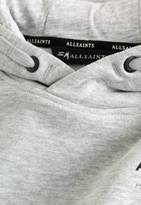 Hoodie grigio con design incrociato, dotato di cordini neri e etichetta con logo sul collo. Tessuto morbido e strutturato.