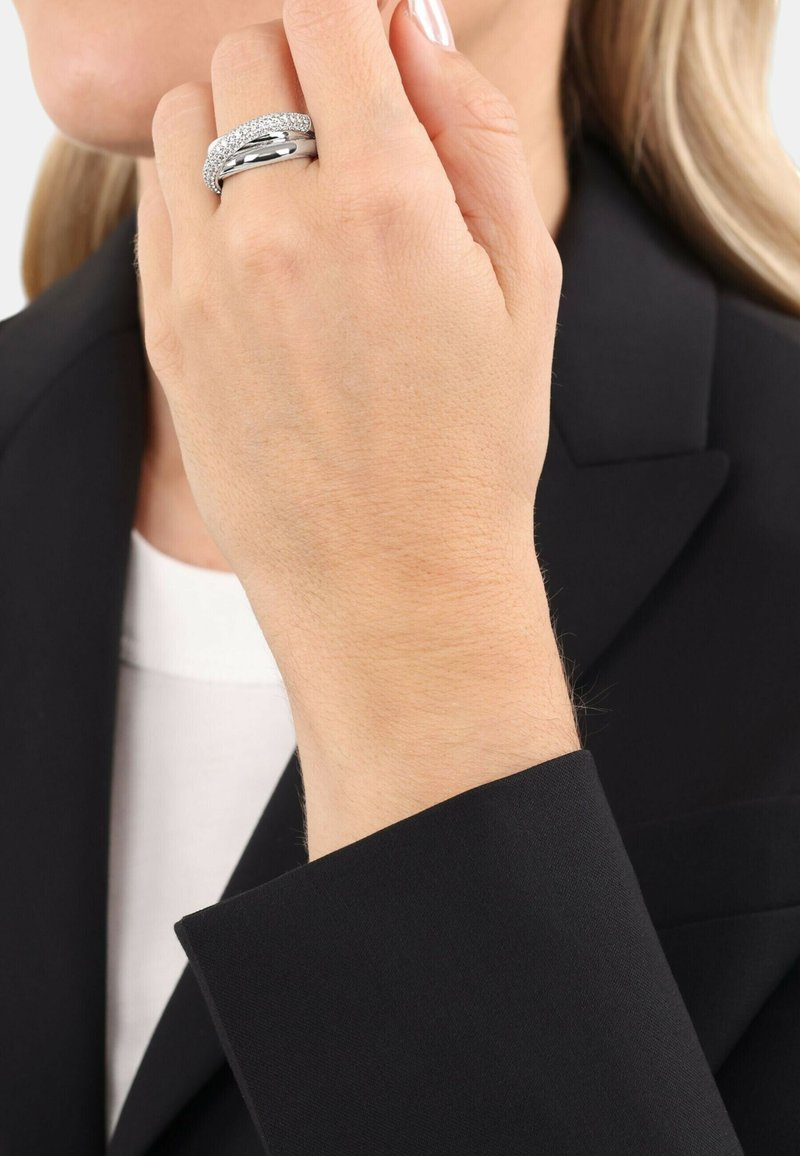 Silverring med en texturerad band, prydd med inslagna klara stenar, bärs på en hand nära kragen av en svart kavaj. Mjuk hudstruktur synlig.