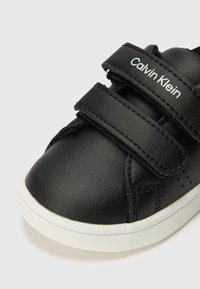Sapatilha preta em couro com sola branca, apresentando duas tiras de velcro e o logotipo "Calvin Klein" em branco na tira superior. Detalhe de superfície texturizada.