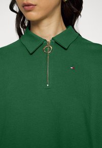 Zelené polo tričko s texturovaným povrchem, s předním zipem a kulatým zlatým táhlem a malou nášivkou v červené, bílé a modré barvě.