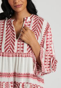 Vit blus med röd geometrisk broderi, med V-ringning, långa utsvängda ärmar och rynkdetalj längs fållen.