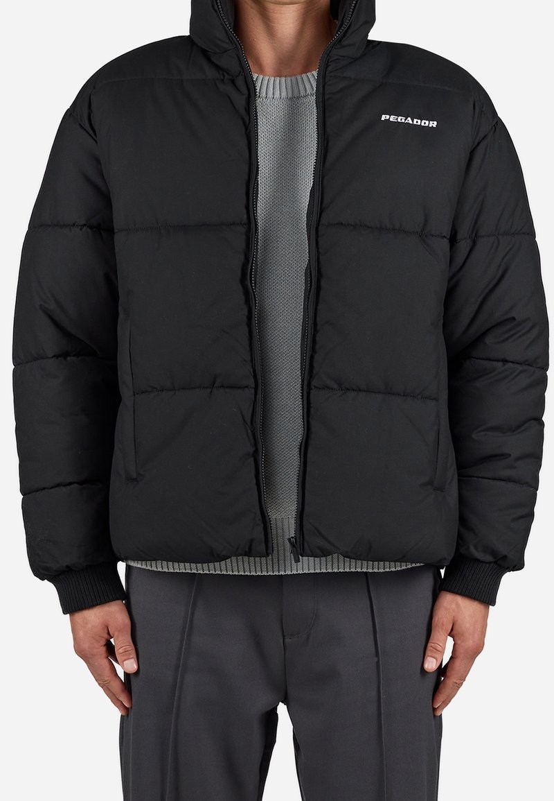 Homme portant une doudoune noire à capuche par-dessus un pull gris en maille et un pantalon gris foncé, debout devant un fond uni.