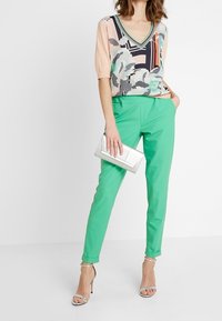 Korte mouwen, V-hals blouse met een bloemen- en geometrisch patroon in pastelkleuren; groene, gepaste broek; zilveren clutch; nude hakken.