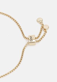 Justerbar gullkjede armbånd med en glidende lås gravert "DKNY" og små runde charm på hvit bakgrunn.