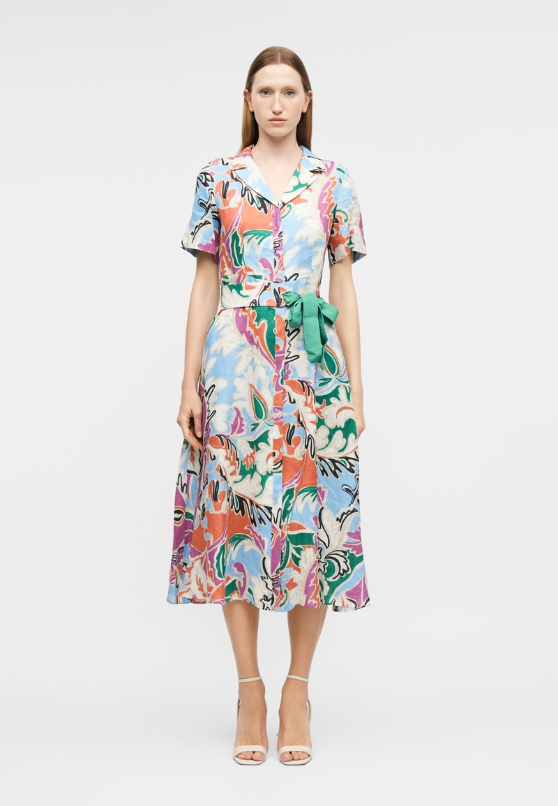 Femme portant une robe midi à imprimé floral abstrait coloré, à manches courtes, col, boutons sur le devant, et une ceinture verte à la taille, associée à des escarpins blancs.