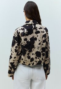 Kort, cropped jacka i beige tyg med svart splattermönster. Har en krage och långa ärmar, som visar en texturerad yta.
