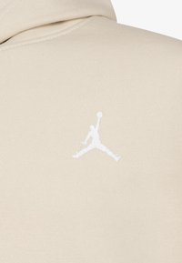 Kreminė gobtuvė su baltu išsiuvinėtu Jumpman logotipu nugarinėje pusėje. Minkšta tekstūra, laisvalaikio stilius su gobtuvu ir raukšlėmis ant rankogalių.
