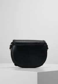 Sac en cuir noir à bandoulière, avec des bords arrondis, fermeture à rabat et sangle réglable, exposé sur un socle blanc devant un fond uni.