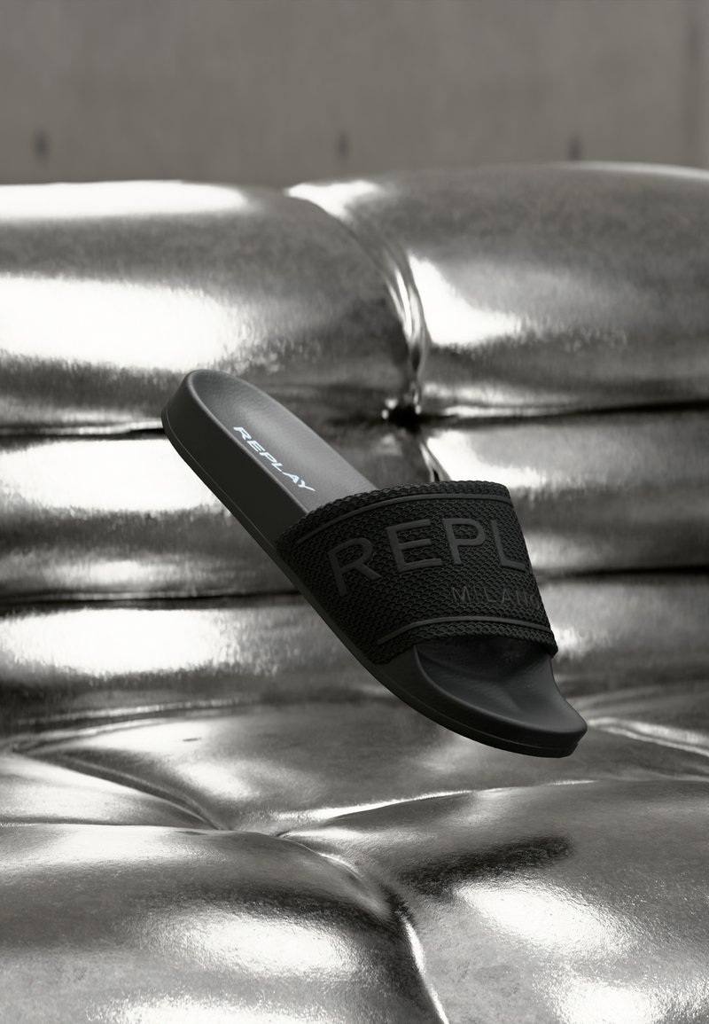 Μαύρο σανδάλι με λουρί με υφή και ανάγλυφο λογότυπο "REPLAY". Κομψός, λιτός σχεδιασμός που ακουμπά σε γυαλιστερή ασημένια επιφάνεια.