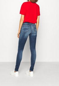 Mujer vestida con unos pantalones ajustados azules desgastados, una camiseta roja de manga corta y zapatillas blancas, de pie de espaldas sobre un fondo blanco liso.