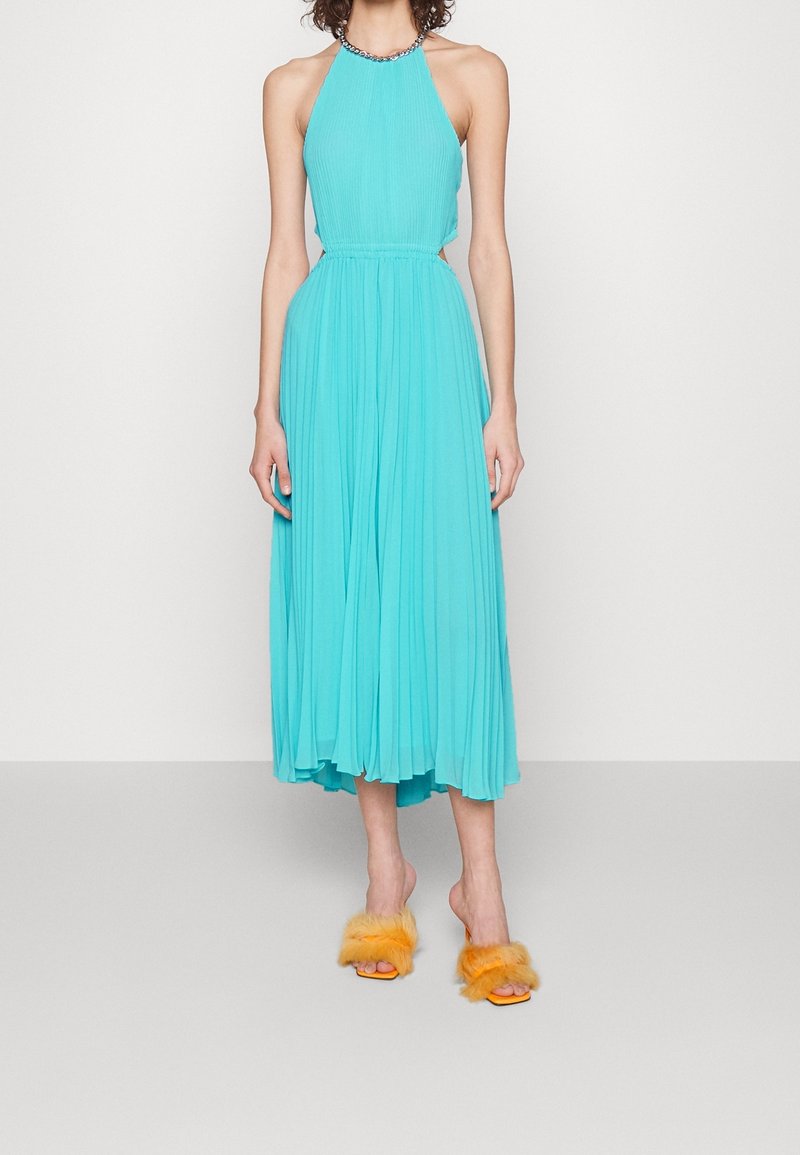 Une robe plissée à col licou d'un turquoise éclatant, avec un corsage ajusté et une jupe fluide. Associée à des sandales slides en fourrure orange.