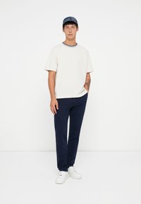 White crew-neck t-shirt with blue trim, paired with navy blue trousers and white sneakers. Model stands against a plain background.