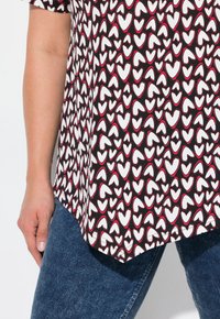 Haut noir avec un motif de cœurs en blanc et bordé de rouge. Le t-shirt a un ourlet arrondi et est associé à un jean bleu.