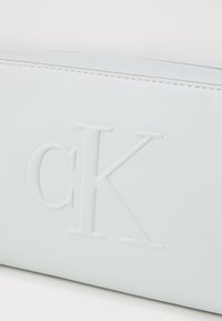 Calvin Klein vit syntetisk läderväska med präglad "CK"-logotyp, slät textur och kontrasterande sömmar längs kanterna.