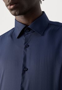 Hombre vestido con una camisa de botón azul oscuro y cuello puntiagudo, mostrado desde el cuello hasta la mitad del pecho sobre un fondo simple.