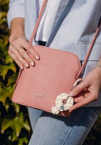 Růžová crossbody taška z texturované kůže se stříbrným logem, s krémově zbarveným přívěskem a tenkým ramenním popruhem, kterou drží osoba.
