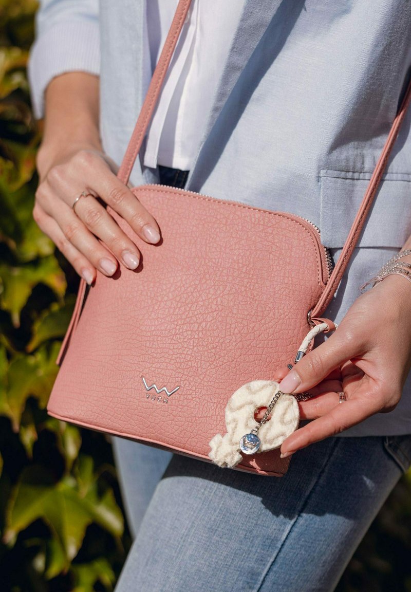 Růžová crossbody taška z texturované kůže se stříbrným logem, s krémově zbarveným přívěskem a tenkým ramenním popruhem, kterou drží osoba.