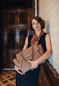 Žena s krátkými hnědými vlasy v černých bezrámových šatech a vzorovaným šátkem, držící velkou hnědou kabelku, stojí uvnitř vedle zdobené zdi.