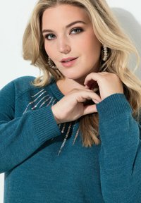 Türkiser Strickpullover mit rundem Ausschnitt und Paillettenverzierungen auf der Brust. Das Modell hat wellige blonde Haare und trägt große Ohrringe.