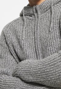 Grå, ribbad stickad huvtröja med zippered hals och dragsko; har en tjock textur och en avslappnad passform, vilket framhäver tygets värme.