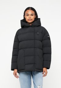 Jachetă puffer neagră cu glugă, având un design capitonat, buzunare laterale și un logo mic pe piept și pe mânecă.