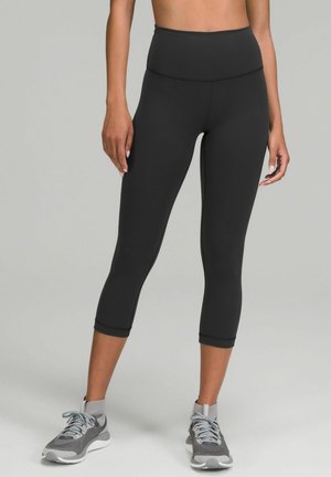 Black high-waisted capri leggings made of stretchy fabric, featuring a smooth finish and ankle-length design paired with gray athletic shoes.