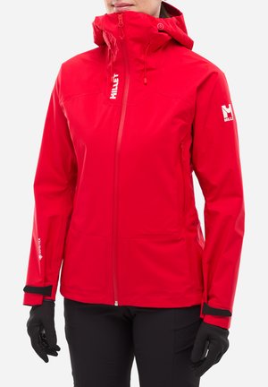 Femme portant une veste imperméable rouge vif Millet avec capuche, gants noirs et pantalon noir, visible de la moitié du torse jusqu'au menton.