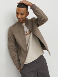 Giubbotto bomber in suede taupe, con chiusura con zip, polsini a costine e una texture liscia. Il modello indossa una camicia beige chiaro sotto.