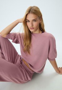 Giovane donna con lunghi capelli biondi, che indossa una felpa color malva e pantaloni della tuta, seduta per terra con un braccio appoggiato sul ginocchio.