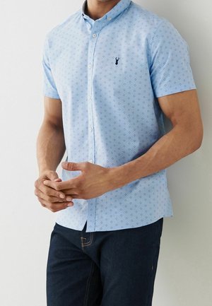 Homme portant une chemise bleu clair à manches courtes avec un petit motif foncé et un jean foncé, debout les mains jointes.
